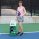 Woman holding a pickleball paddle on a court next to a pickleball ball machine.